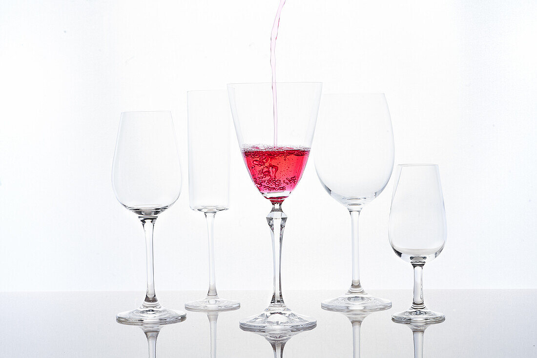 Anonymous person pouring red sparkling wine into goblet laced on mirrored table near various shaped glasses against white background