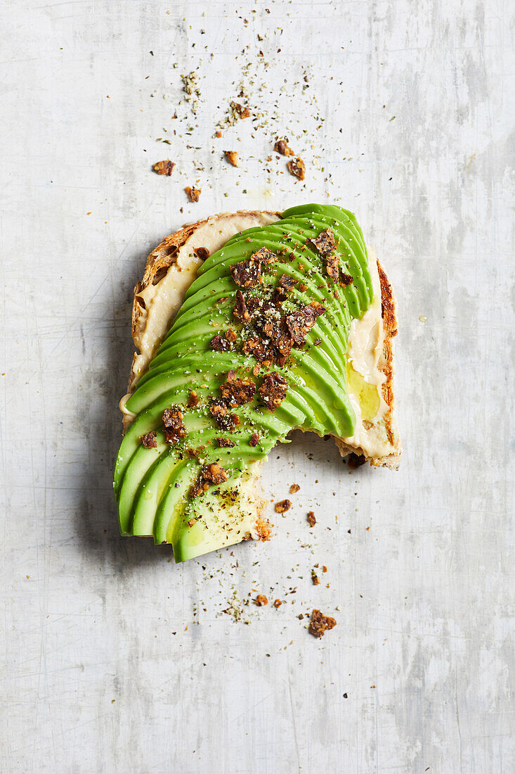 Blick von oben auf einen angebissenen Toast mit Hummus und Avocado, serviert auf einem Marmortisch in der Küche