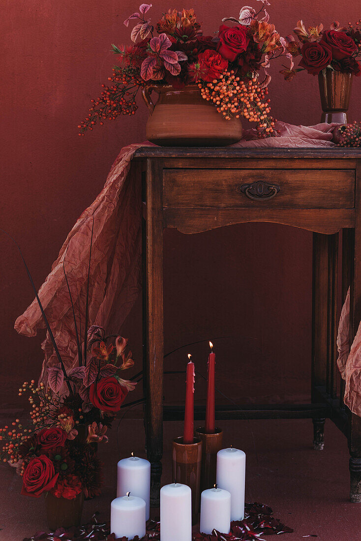 Wooden old fashioned table decorated with fabric and vases of red flowers near burning candles in studio