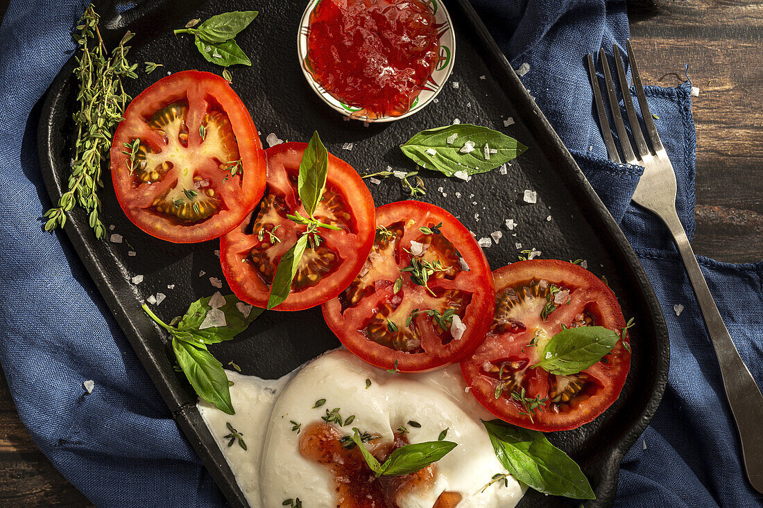 From above of appetizing burrata cheese with sauce and slices of fresh tomato with thyme and basil leaves served on cast iron tray placed on table cloth on wooden table with fork