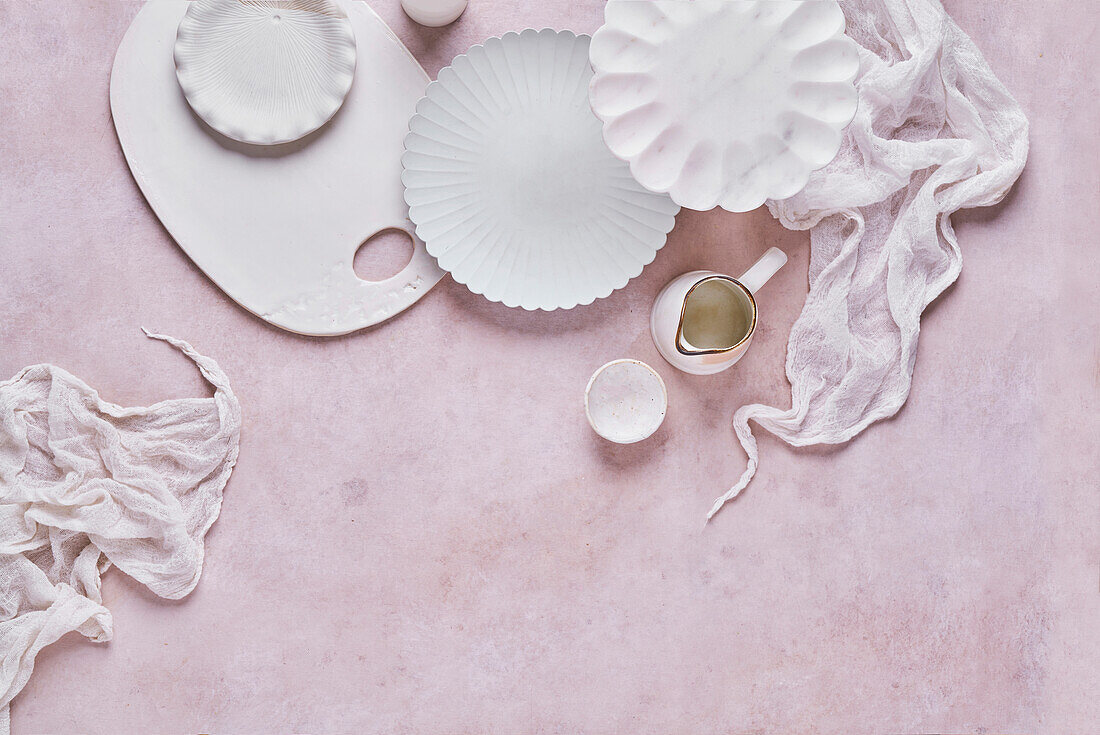 Top view set of various white ceramic plates with cutting board and small pitcher placed on pale pink surface with fabric