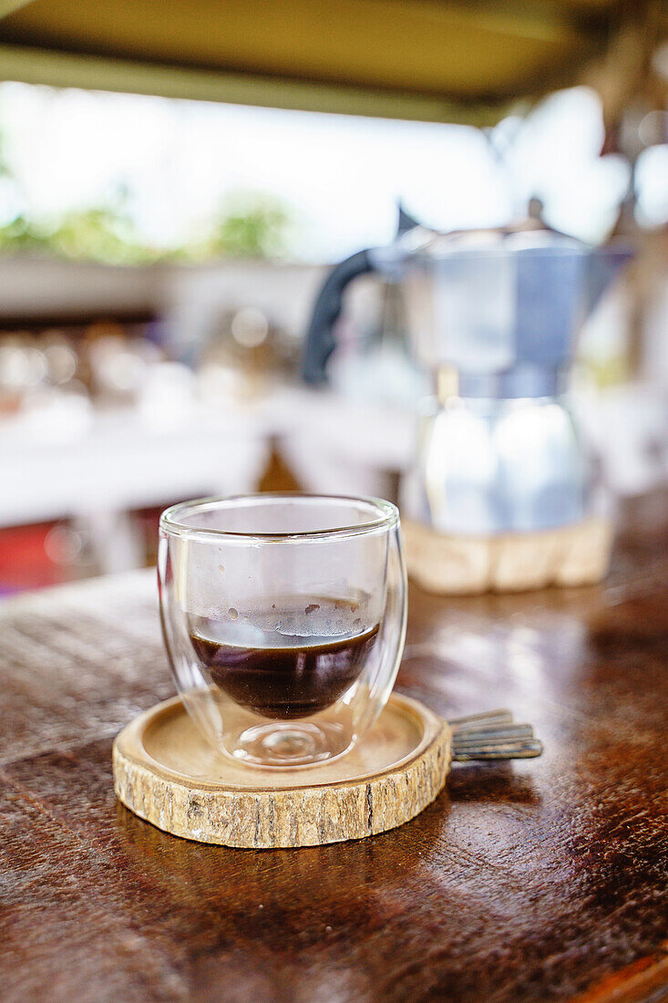 Ein Glas frisches Heißgetränk aus einer Moka-Kanne auf einem Holztresen in einem Kaffeehaus