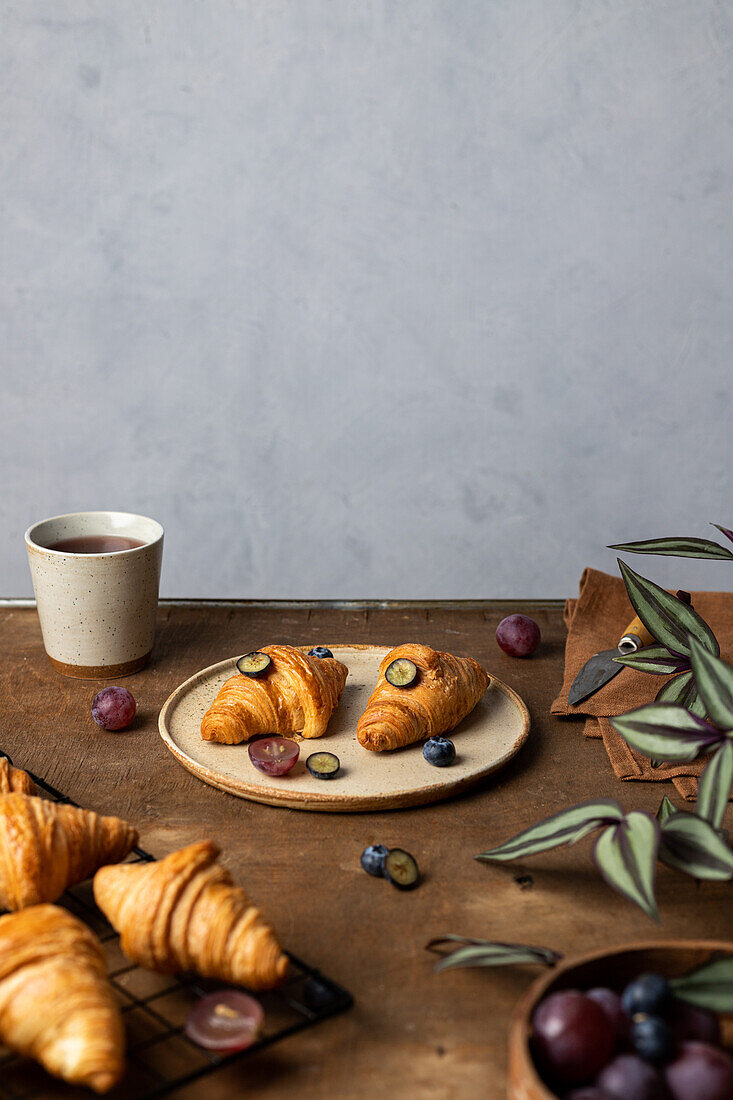 Leckere frisch gebackene Croissants auf einem Teller mit Früchten neben einer Tasse Tee auf einem Holztisch am Morgen in einem hellen Raum
