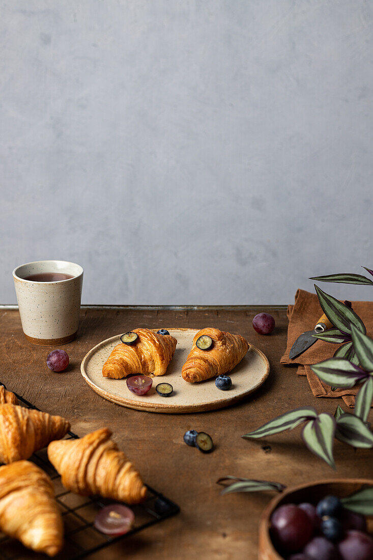 Tasty fresh baked croissants served on plate with fruits placed near cup of tea on wooden table in morning time in light room