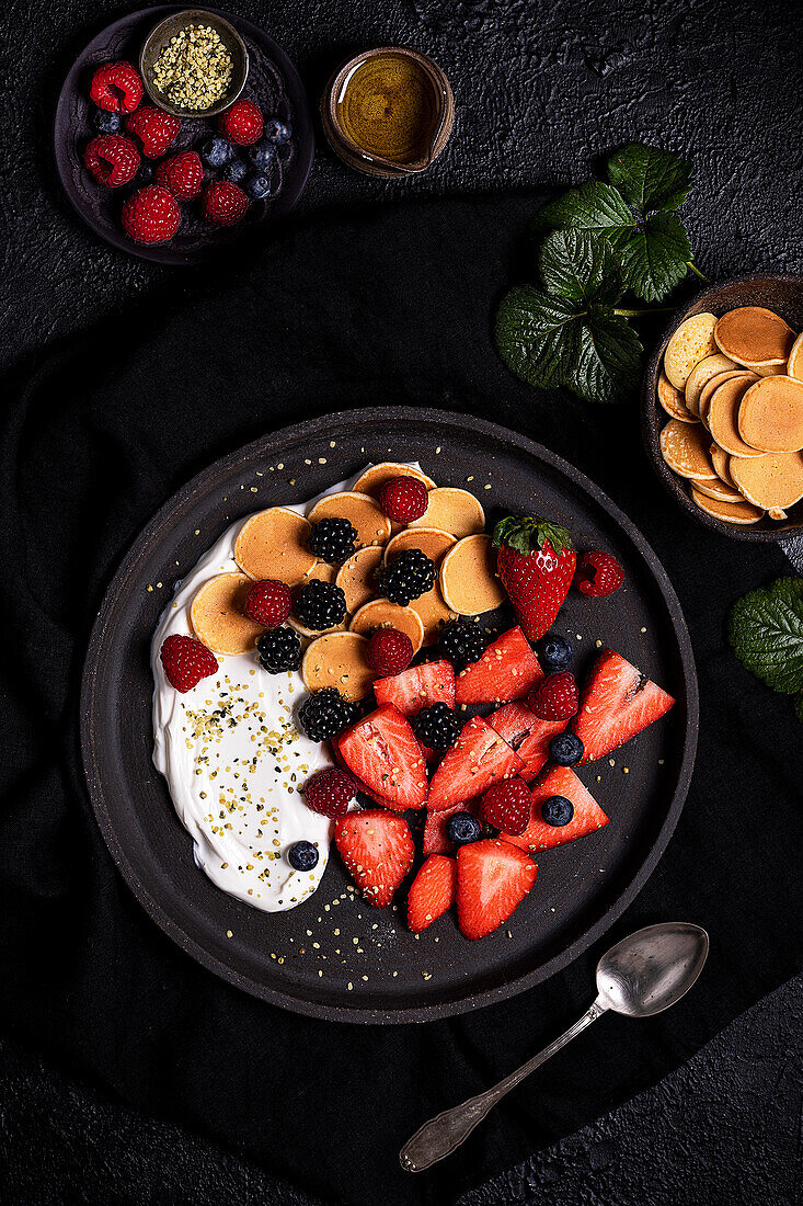 Ansicht von oben auf ein leckeres, gesundes, hausgemachtes Frühstück mit frischen, verschiedenen Beeren, serviert mit Joghurt und Keksen in einem schwarzen Teller auf einem Tisch mit Zutaten