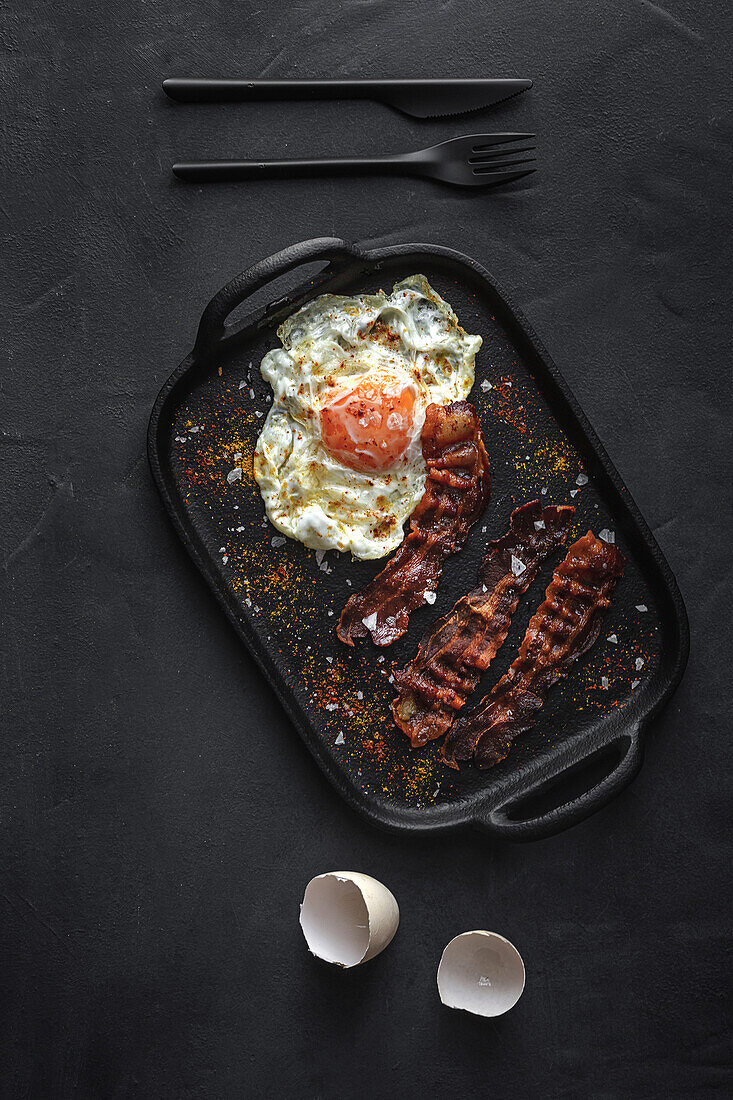 Top view of sunny side up egg with fried bacon slices and condiments on tray against cutlery on dark background