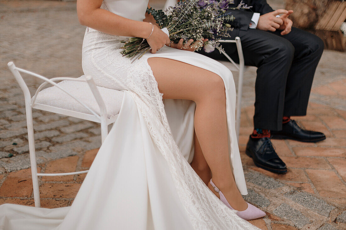 Anonyme Braut in einem eleganten weißen Maxikleid, das die Beine freigibt, hält einen Strauß zarter Blumen und sitzt neben dem Bräutigam im schwarzen Anzug während der Hochzeitsfeier