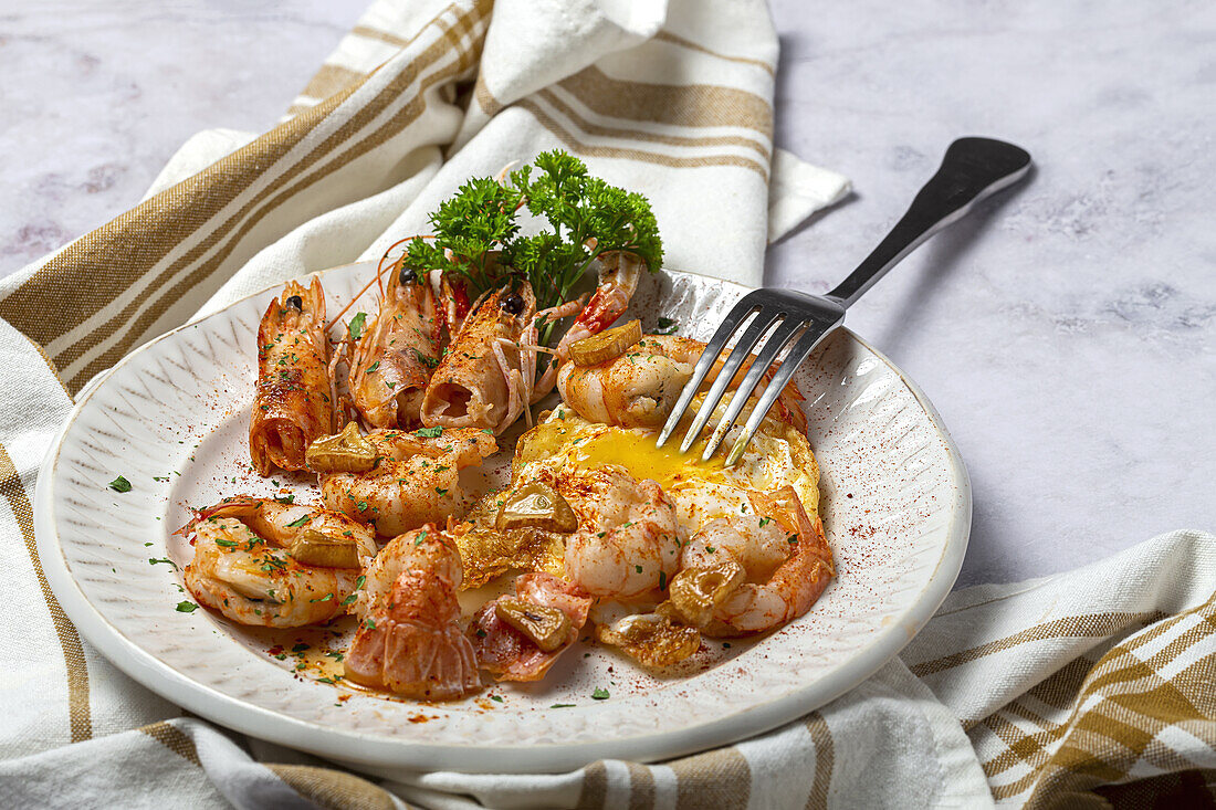 From above appetizing fried eggs with king prawns served on white plate with green herbs and seasoning on towel in light kitchen