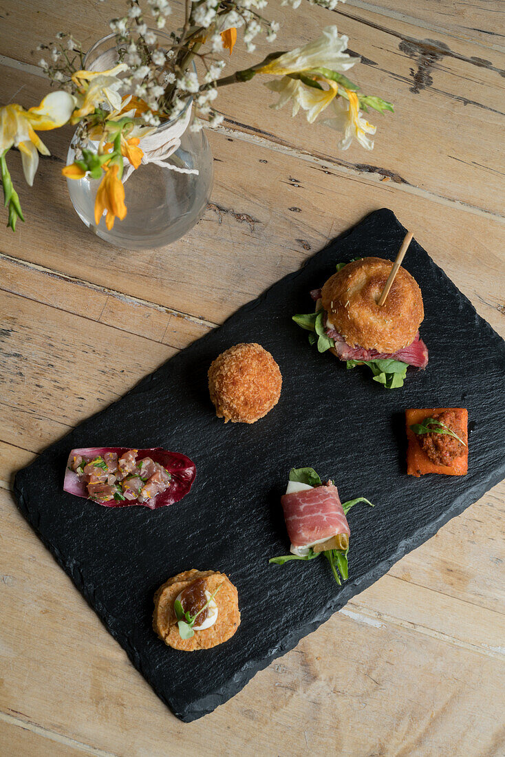 Top view of assorted delicious appetizers served on black board near glass vase with blooming bouquet placed on wooden surface
