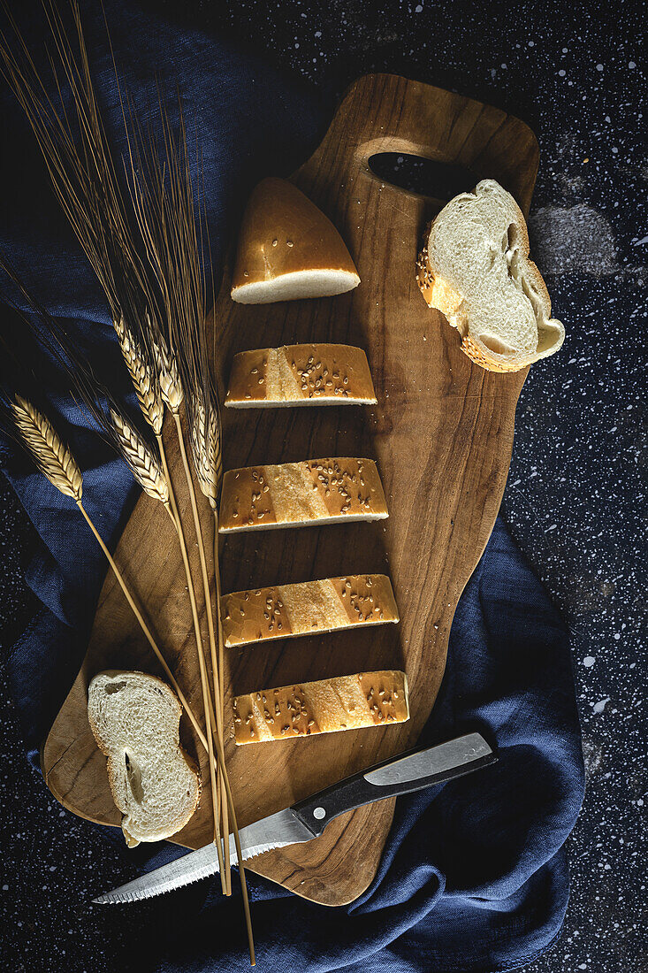 Ansicht von oben auf Stücke von Weißbrot neben Messer und Weizenspitzen auf Holzbrett