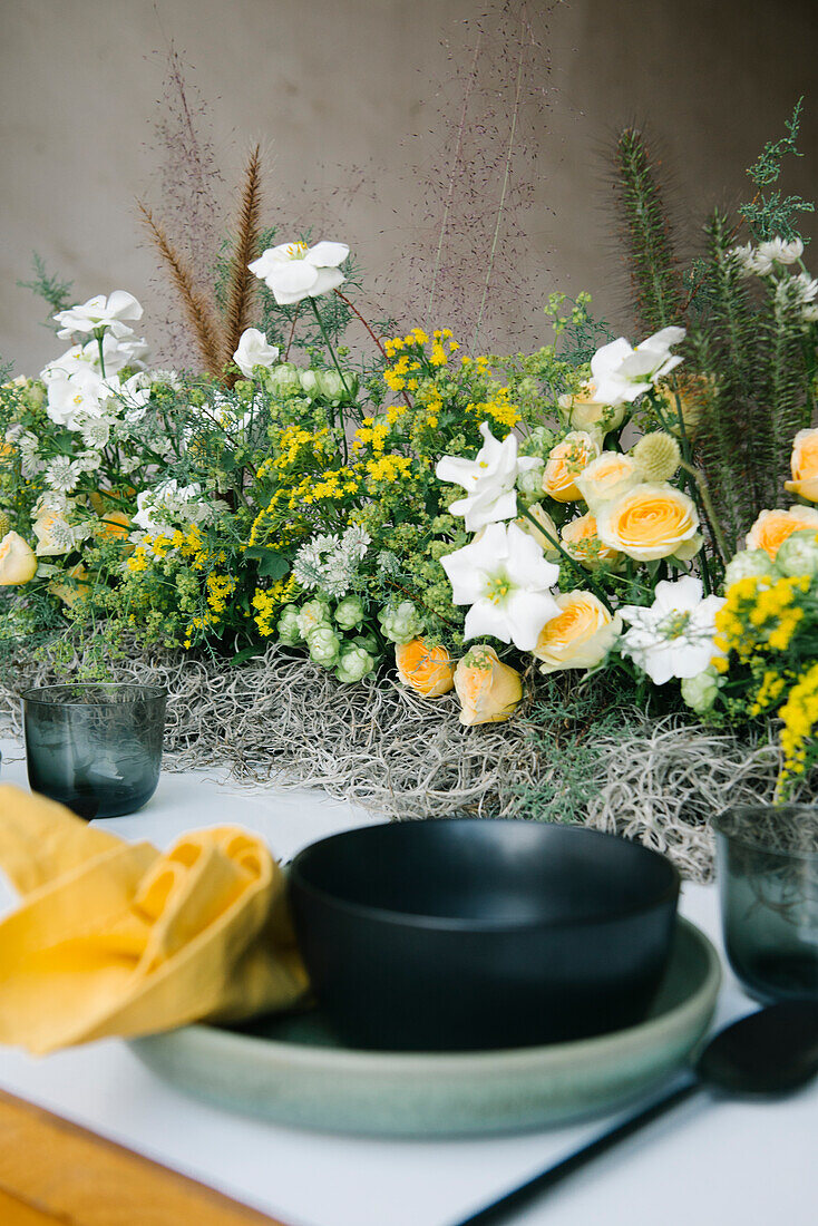 Blick von oben auf einen Teller und eine Schüssel mit Besteck und Serviette, die auf einem mit zarten frischen weißen und gelben Blumen dekorierten Tisch stehen