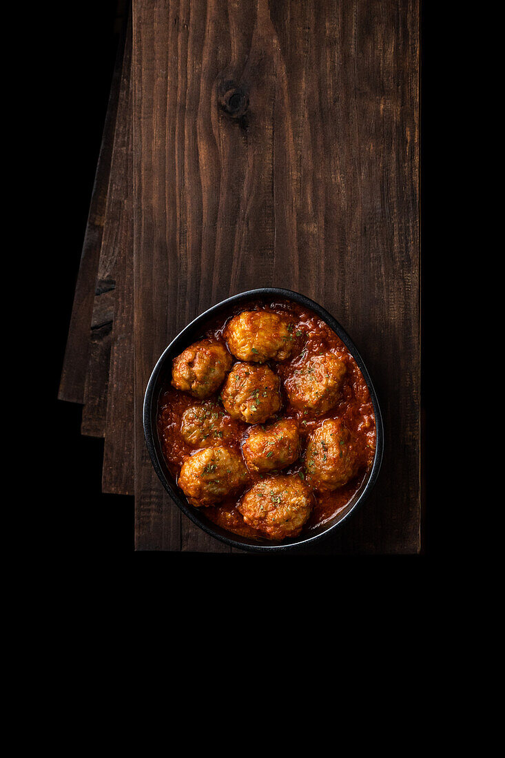 Draufsicht auf eine schwarze Schüssel mit leckeren Fleischbällchen in Tomatensauce mit Kräutern, die auf einem Stapel Holzbretter in einem dunklen Raum platziert sind