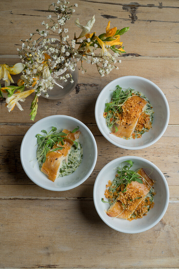 Draufsicht auf appetitlichen Fisch mit Soße und Grünzeug in Keramikschalen auf Holzboden neben Glasvase mit blühendem Blumenstrauß