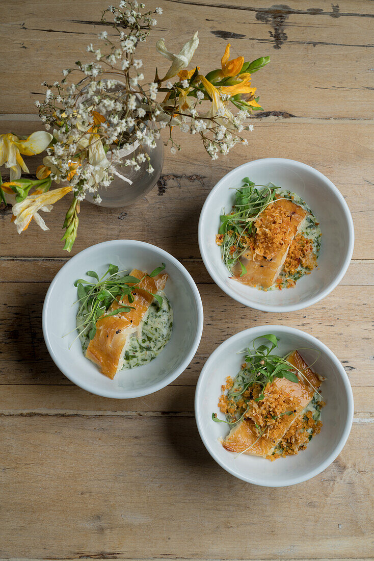 Top view of appetizing fish served with sauce and greens in ceramic bowls placed on wooden floor near glass vase with blooming bouquet