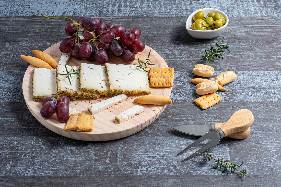Von oben appetitlicher Käse serviert auf Holztisch mit reifen Trauben und Crackern, dekoriert mit Rosmarinzweigen neben Oliven in Schalen auf dem Tisch