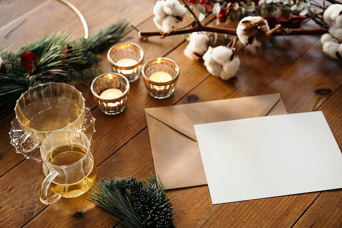 From above of Christmas composition with burning candles and cups of tea placed near empty postcard on wooden table decorated with fir branches and cotton twig