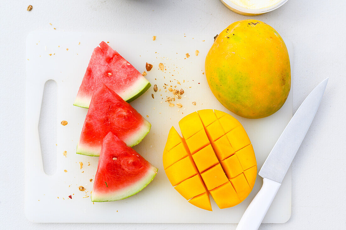 Draufsicht auf frische, in Scheiben geschnittene Wassermelone und orangefarbene, saftige, halbierte Mango, die mit einem Messer auf ein weißes Schneidebrett gelegt wird