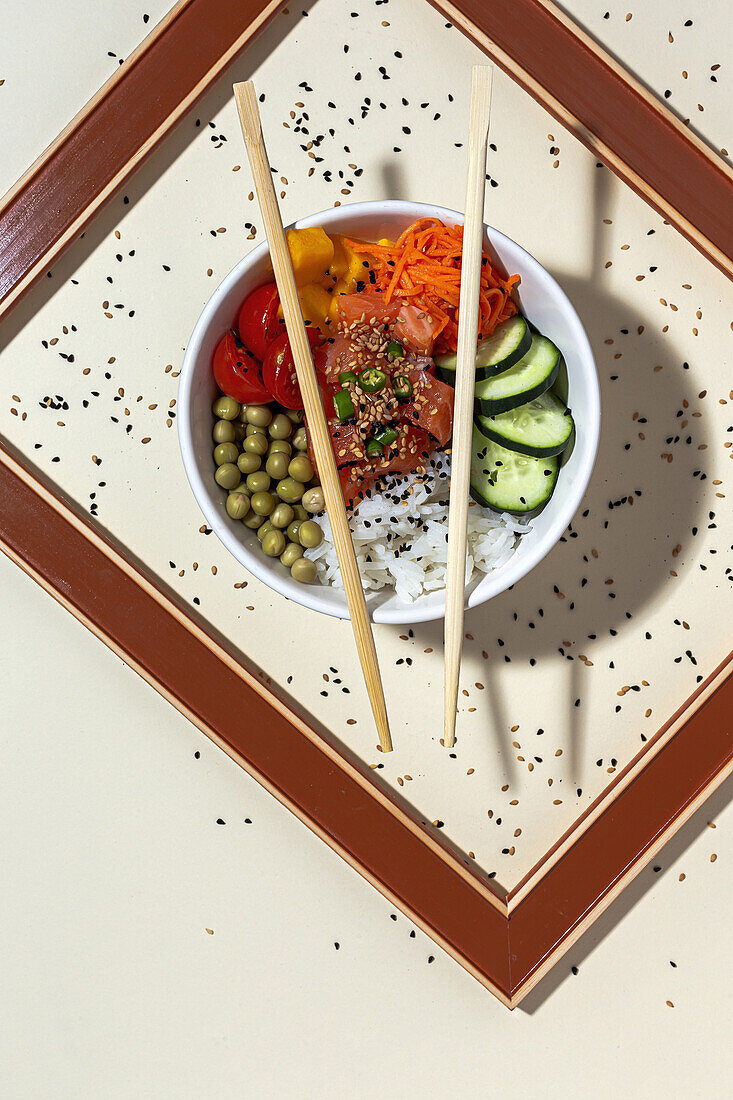 From above white bowl with tasty poke dish and chopsticks placed behind frame on table covered with sesame seeds