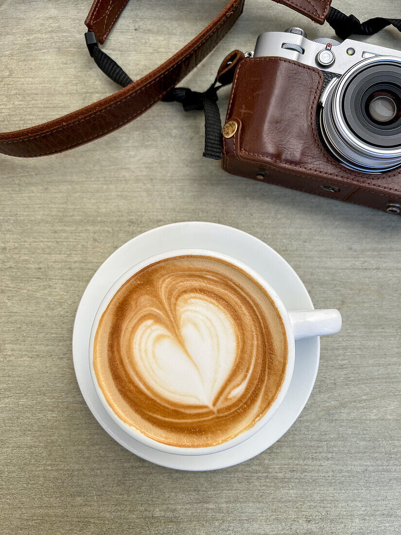 Draufsicht auf einen Becher mit heißem Kaffee mit gezeichnetem Herz, der auf einem Tisch neben einer alten Fotokamera in einer braunen Ledertasche steht