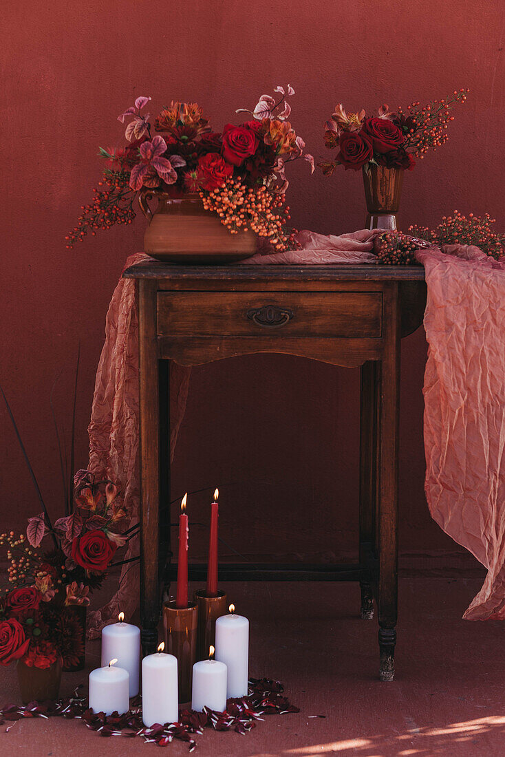 Altmodischer Holztisch, dekoriert mit Stoff und Vasen mit roten Blumen neben brennenden Kerzen in einem Atelier