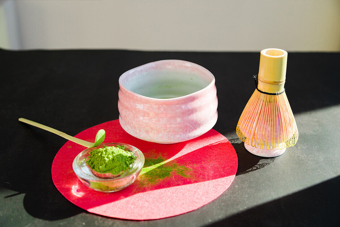 Blick von oben auf pulverisierten Matcha-Tee mit Bambus-Chashaku neben Keramikschale und Chasen-Besen auf schwarzem Tisch