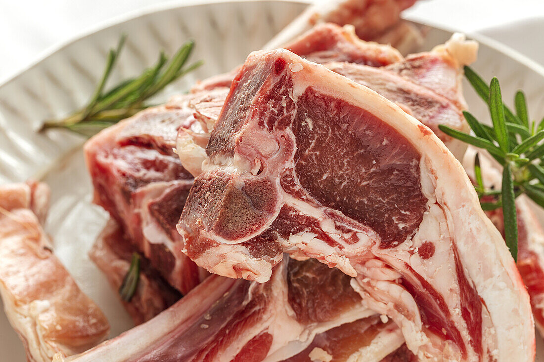 From above of uncooked lamb chops served on white ceramic plate with fresh green rosemary on table in light kitchen