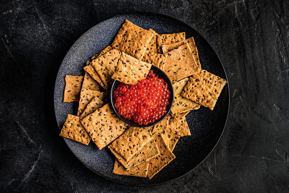From above red fresh trout fish caviar in a bowl served with sesame seed crackers on concrete table background
