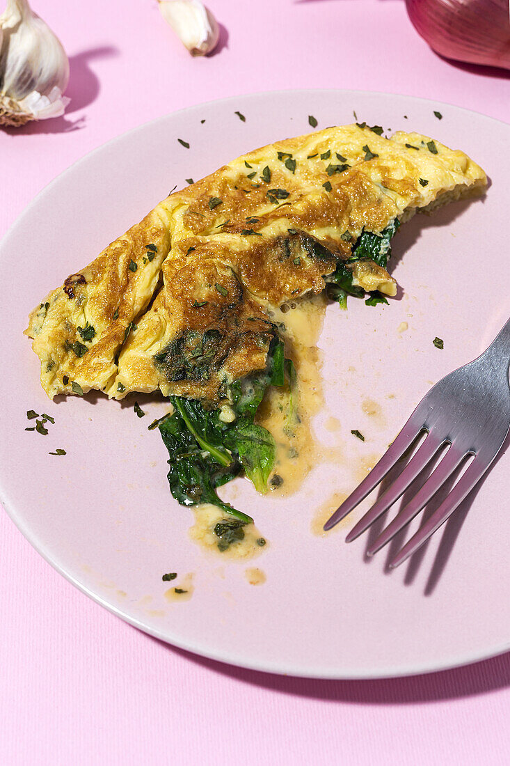 Tasty omelette on plate against fresh parsley sprigs and red onion with garlic cloves on pink background