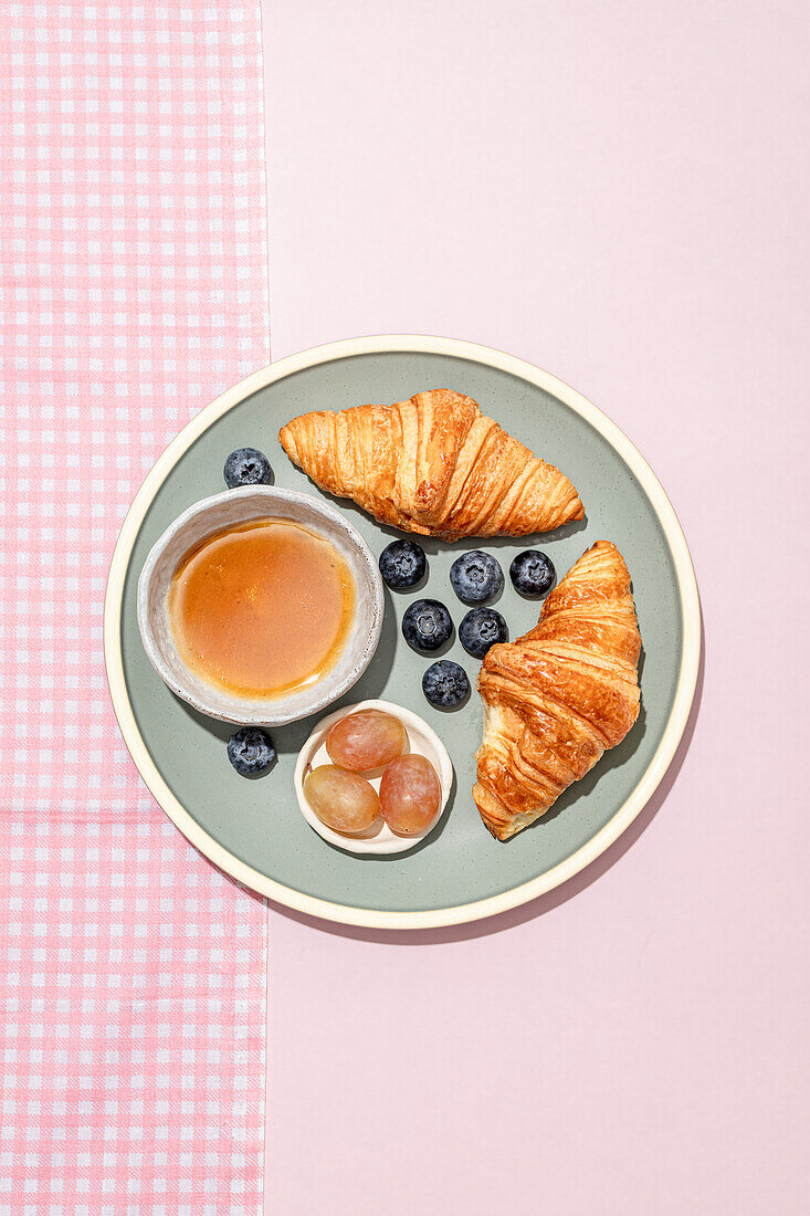 Draufsicht auf leckere Croissants auf Keramikteller mit frischen Blaubeeren und Marmelade auf dem rosa Tisch