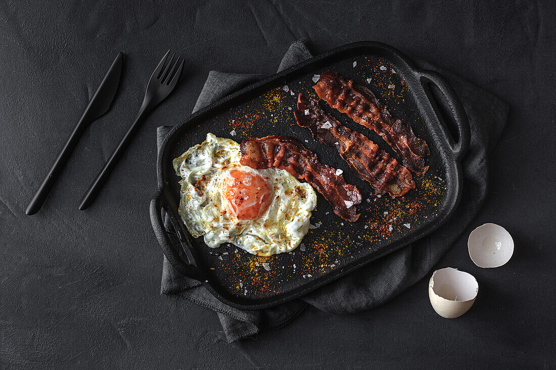 Top view of sunny side up egg with fried bacon slices and condiments on tray against cutlery on dark background