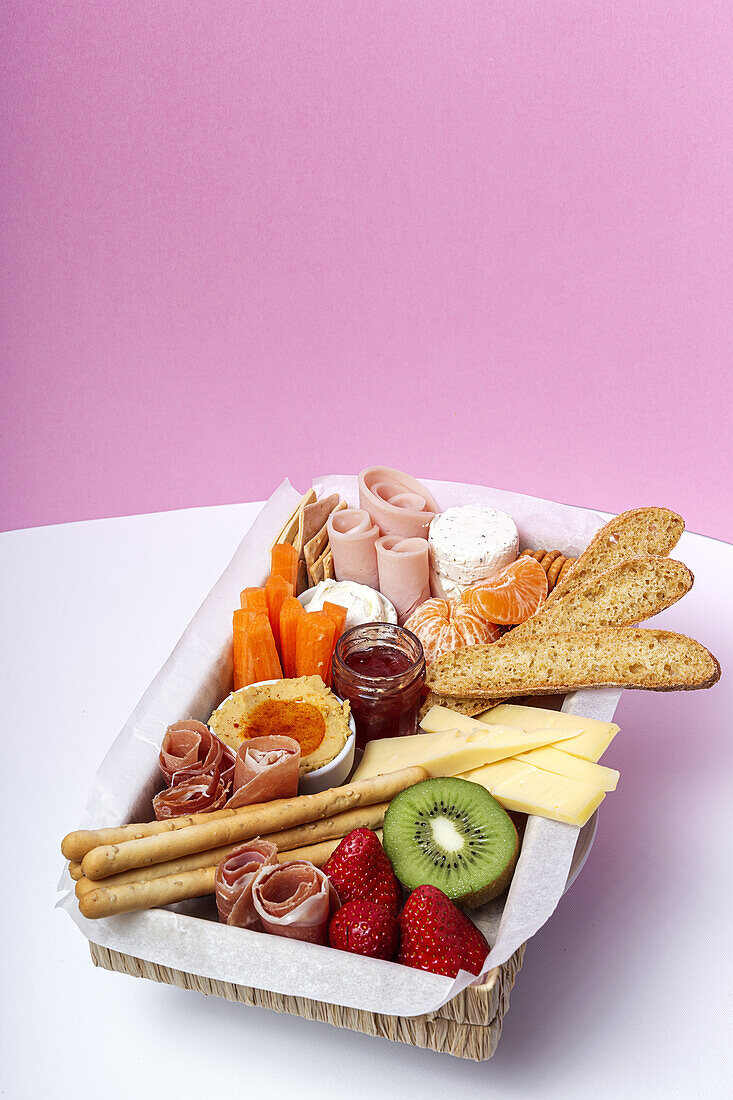 From above brunch box with assorted sliced meats various types of cheese and crispbreads arranged near ripe cup kiwi sweet strawberries and peeled mandarin near jam in glass jar on colorful background