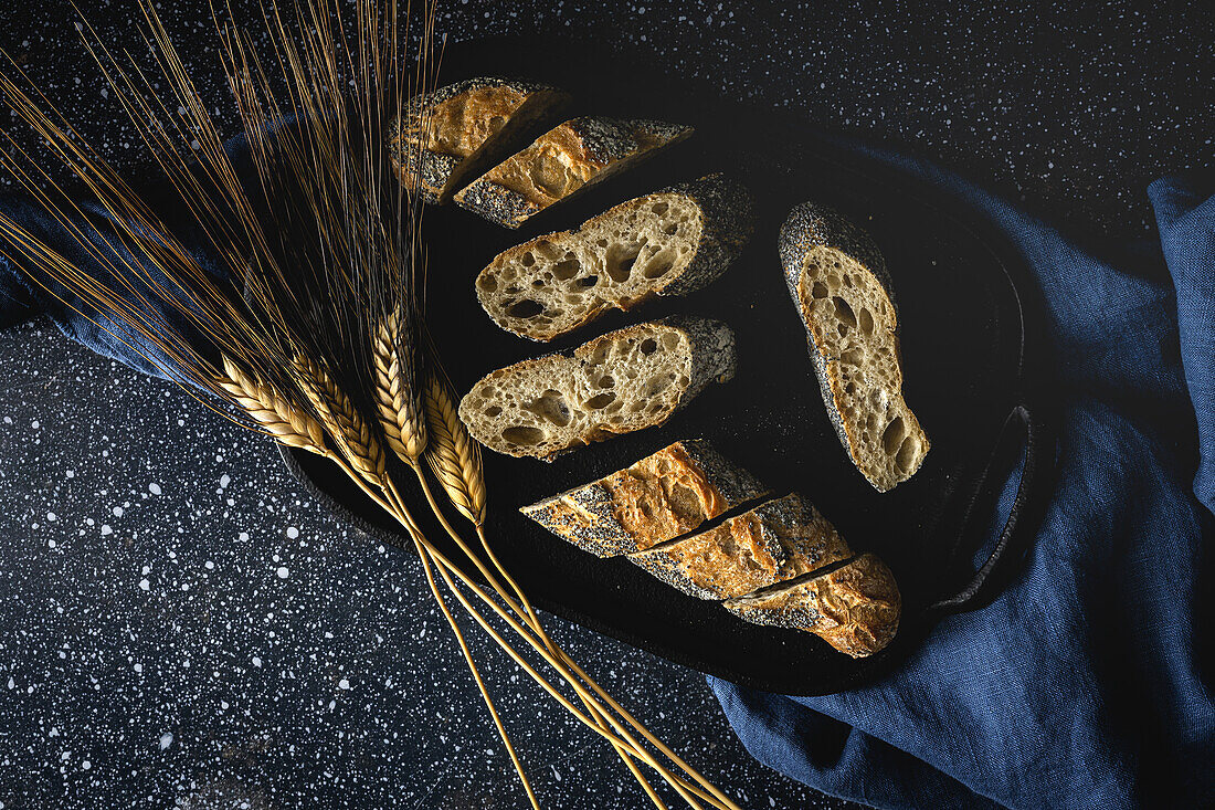 From above of appetizing crusty bread near wheat spikes and dark fabric on table