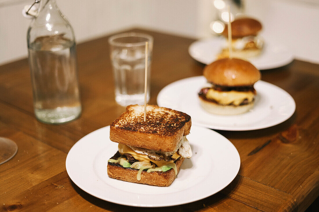 Blick von oben auf ein appetitliches Sandwich mit gebratenem Toast, das auf einem weißen Teller liegt und mit einem Zahnstocher durchbohrt wird, während es auf einem Holztisch neben einer Flasche Wasser serviert wird