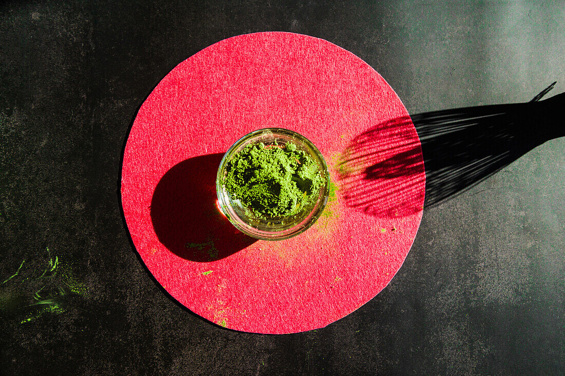 Top view of glass bowl with dried matcha tea on round bright circle on black background with shadow of bamboo whisk