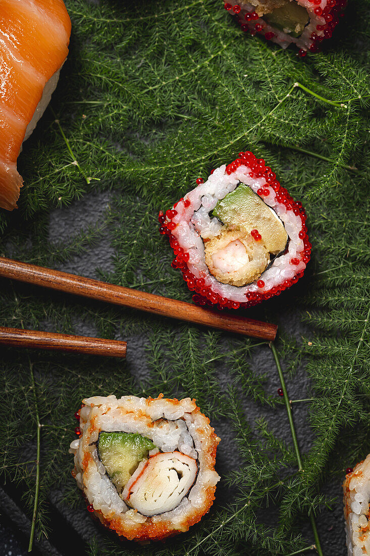 Tasty fresh assorted sushi served on green plant twigs on black plate with soy sauce on marble table near chopsticks