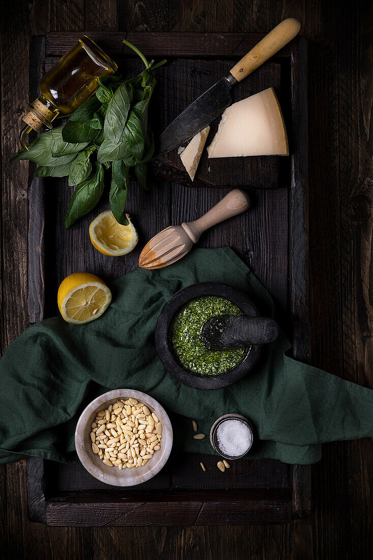 Komposition von oben mit Zutaten für eine traditionelle Pesto-Soße, darunter Parmesankäse, Basilikumblätter, Zitrone und Pinienkerne, auf einem dunklen Tisch