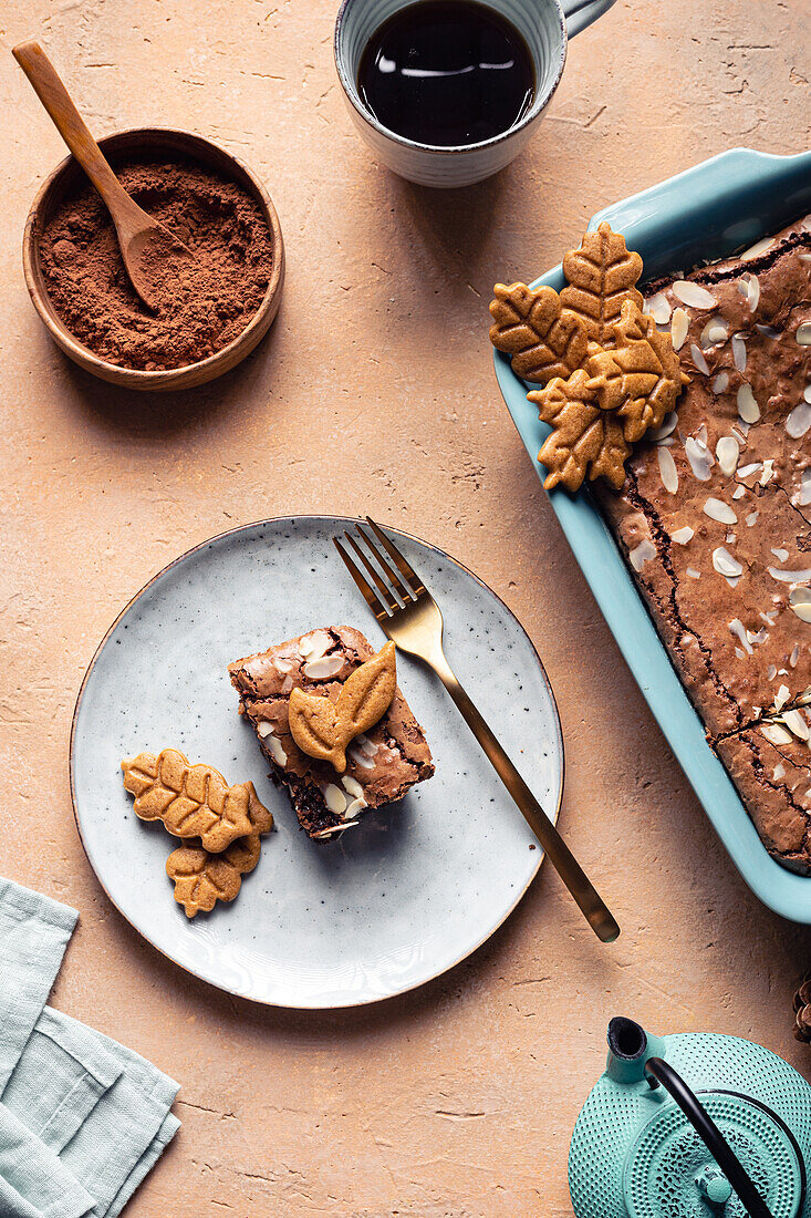 Ein Stück süßer, frisch gebackener Schokoladen-Brownie mit Keksen, serviert auf einem Teller neben einer Backform auf einem Tisch mit einer Tasse Kaffee