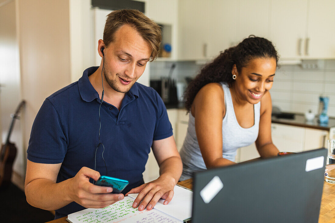 Smiling young multiracial couple using gadgets and devices while sitting at counter in kitchen and studying