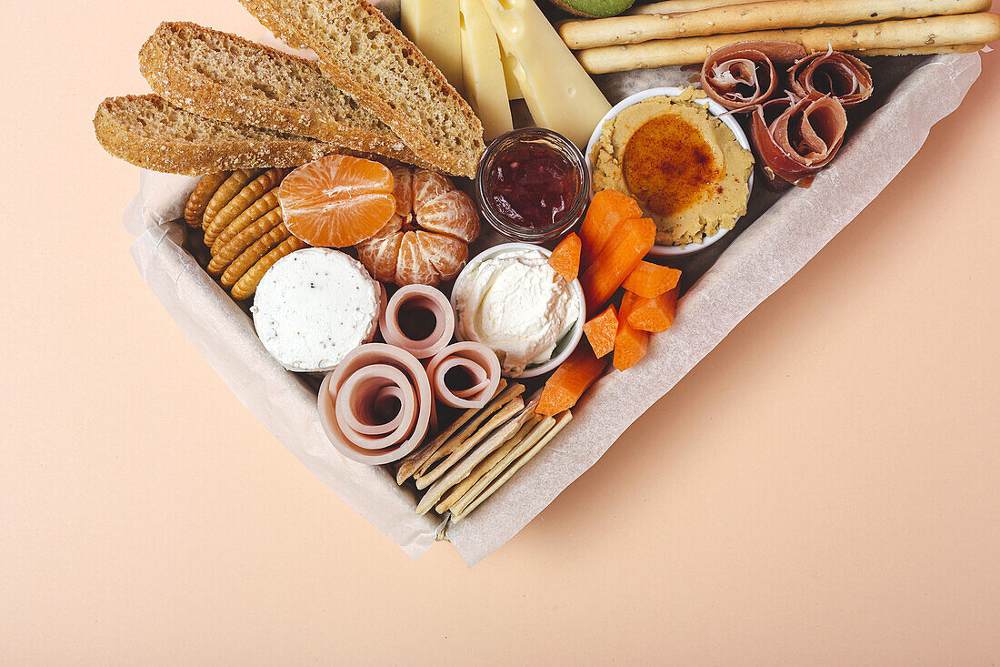 Von oben Brunch-Box mit verschiedenen Wurst- und Käsesorten und Knäckebrot in der Nähe von reifen Kiwis, süßen Erdbeeren und geschälten Mandarinen in der Nähe von Marmelade in einem Glas auf einem Holztisch in der Nähe einer Serviette