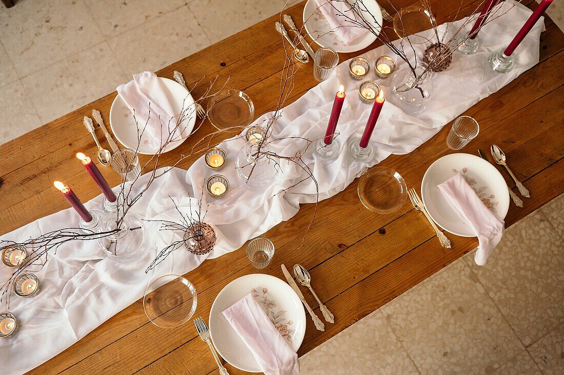 Weißes Tischtuch und Teller auf einem festlich gedeckten Tisch mit brennenden Kerzen und trockenen Baumzweigen von oben