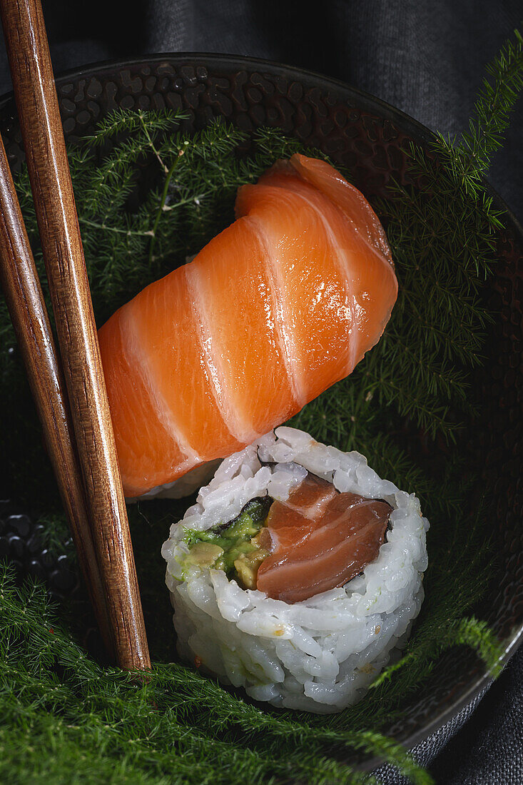 Leckeres frisches Sushi auf grünen Pflanzenzweigen auf schwarzem Teller mit Sojasauce auf Marmortisch neben Stäbchen