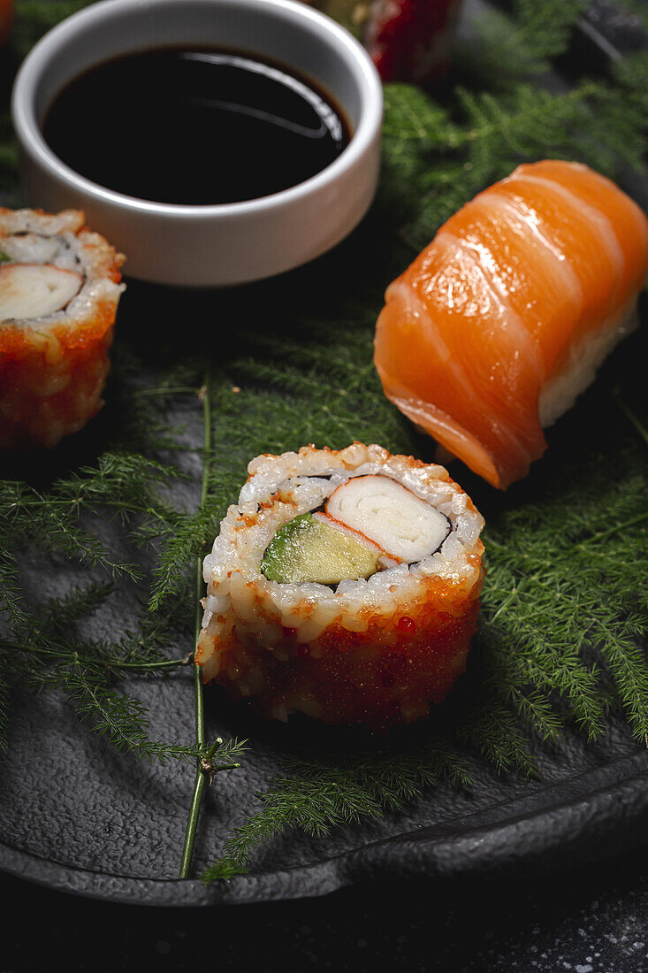 Tasty fresh assorted sushi served on green plant twigs on black plate with soy sauce on marble table