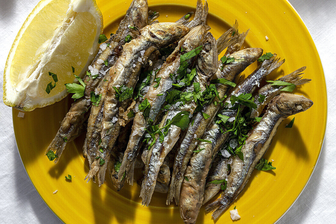 Top view plate of appetizing grilled anchovies served on table with piece of lemon and fresh herbs in restaurant