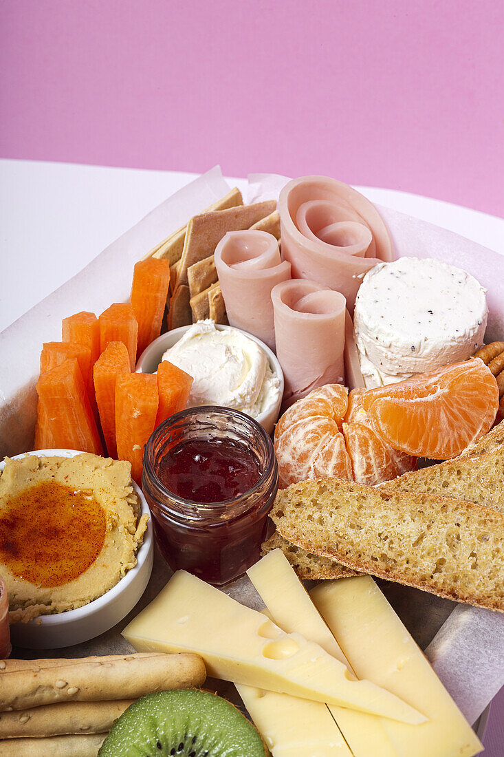 Von oben Brunch-Box mit verschiedenen Wurst- und Käsesorten und Knäckebrot in der Nähe von reifen Kiwis, süßen Erdbeeren und geschälten Mandarinen in der Nähe von Marmelade in einem Glas auf einem Holztisch in der Nähe einer Serviette