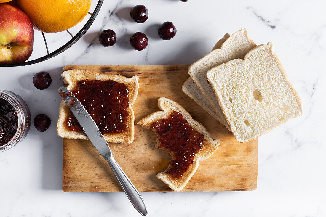 High angle of appetizing crispy toasts with cherry jam served on wooden chopping board near fresh ripe cherries on table