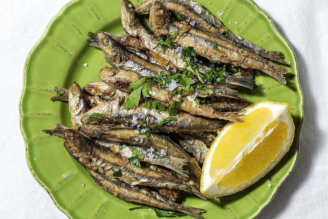 Top view plate of appetizing grilled anchovies served on table with piece of lemon and fresh herbs in restaurant