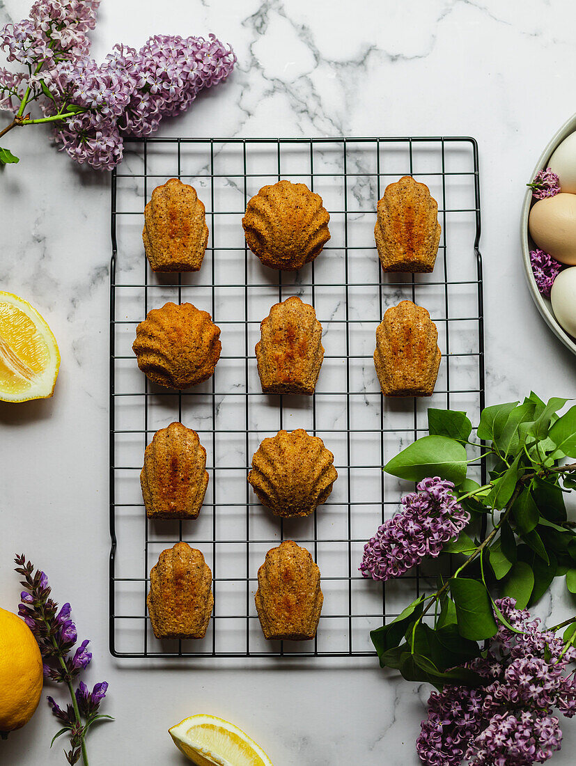 Top view of tasty madeleines on cooling rack near plate with eggs and lavender flowers on marble surface
