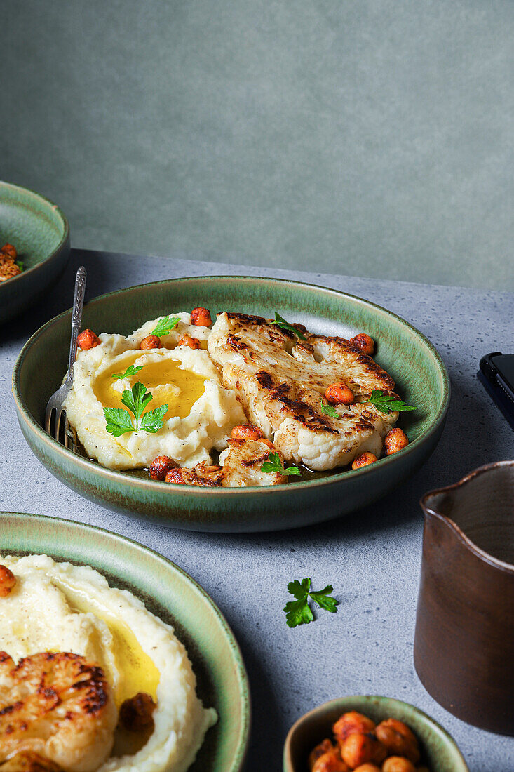 Köstlicher gebratener Blumenkohl mit Kartoffelpüree und gewürzten Harissa-Kichererbsen, serviert auf einem Keramikteller auf grauem Tischhintergrund