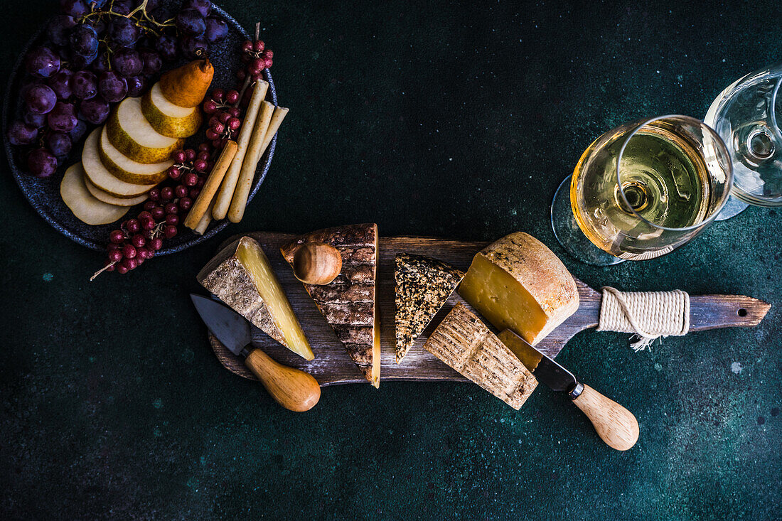 Draufsicht auf Käsesorten auf einem hölzernen Schneidebrett, Obstteller und Weingläser