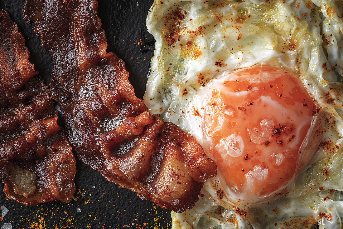 From above of sunny side up egg with fried bacon slices and condiments on dark tray